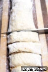 A knife slices through a cylindrical piece of dough, destined to become cinnamon roll muffins, on a wooden surface. The lightly floured dough and the flour-dusted surface hint at the sweet transformation awaiting these evenly cut sections.