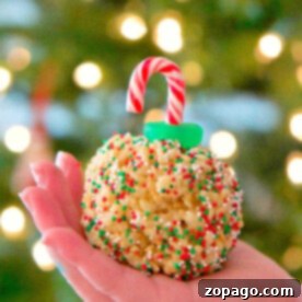 Krispy Holiday Tree Treats 2 A hand holds a festive Rice Krispy treat ornament adorned with sprinkles and a candy cane hook, set against the soft glow of a blurred tree.