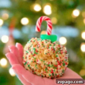 A hand holds a festive Rice Krispy treat ornament adorned with sprinkles and a candy cane hook, set against the soft glow of a blurred Christmas tree.