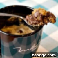 A spoon filled with chocolate chip cookie dough hovers over a black mug, invitingly set on a checkered tablecloth, promising the perfect chocolate chip cookie in a mug.
