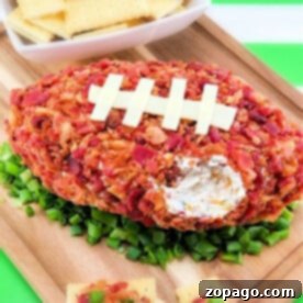 Touchdown Cream Cheese Football Dip 3 A football-shaped cheese ball, perfectly covered in crispy bacon, rests on a bed of fresh greens with white crackers elegantly arranged on a wooden board.