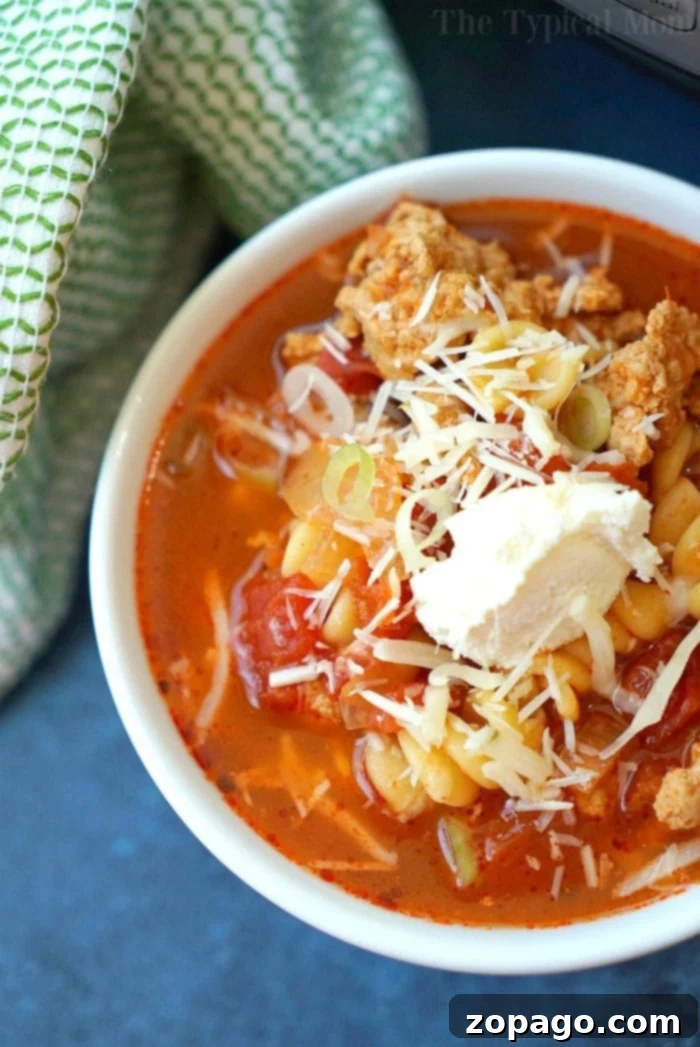 This easy Instant Pot lasagna soup is amazing and healthy!! You can make it in the slow cooker too but in your pressure cooker it's done in just 8 minutes! Your favorite pasta dinner is now a soup and much easier to make. #lasagnasoup #lasagna #soup #instantpot #pressurecooker #easy A close-up of a bowl of minestrone soup garnished with grated cheese, a dollop of cream, and crumbled pieces of bread. The soup contains pasta, tomatoes, and broth reminiscent of instant pot lasagna soup. A green and white cloth appears in the background.