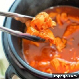 Tongs grasping tender Instant Pot shredded chicken in a pressure cooker brimming with rich red sauce.