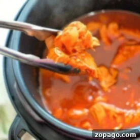 Tongs grasping tender instant pot shredded chicken in a pressure cooker brimming with rich red sauce.