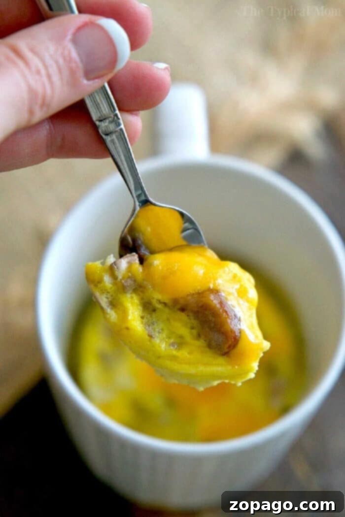 Discover how to make a delicious and fluffy omelette in a mug in just 1 minute using your microwave! This protein-packed, cheesy breakfast is incredibly easy, customizable, and perfect for busy mornings. Kids love these quick scrambled eggs in a cup! A hand holds a spoon with cheesy egg and sausage above a white mug filled with a microwave omelette in a mug.
