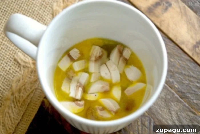 This omelette in a mug recipe is cooked quickly in the microwave and tastes absolutely amazing! It's the perfect breakfast in a mug recipe, packed with protein, and even my kids love it! A white mug filled with a cheesy omelette and sliced mushrooms, representing a delicious omelette in a mug, resting on a wooden surface.