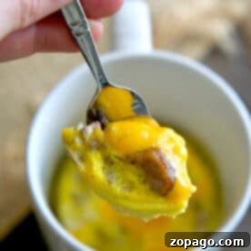 A hand holds a spoon with cheesy egg and sausage above a white mug filled with omelette in a mug.