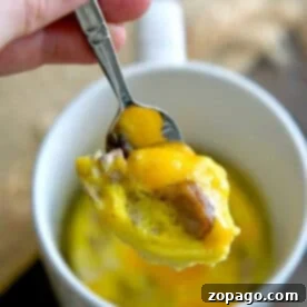 A hand holds a spoon with cheesy egg and sausage above a white mug filled with omelette in a mug.