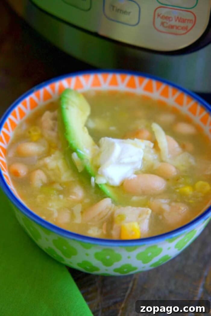 A vibrant bowl of Instant Pot white chicken chili, generously topped with fresh avocado slices and a swirl of cool sour cream, positioned invitingly in front of an electric pressure cooker.
