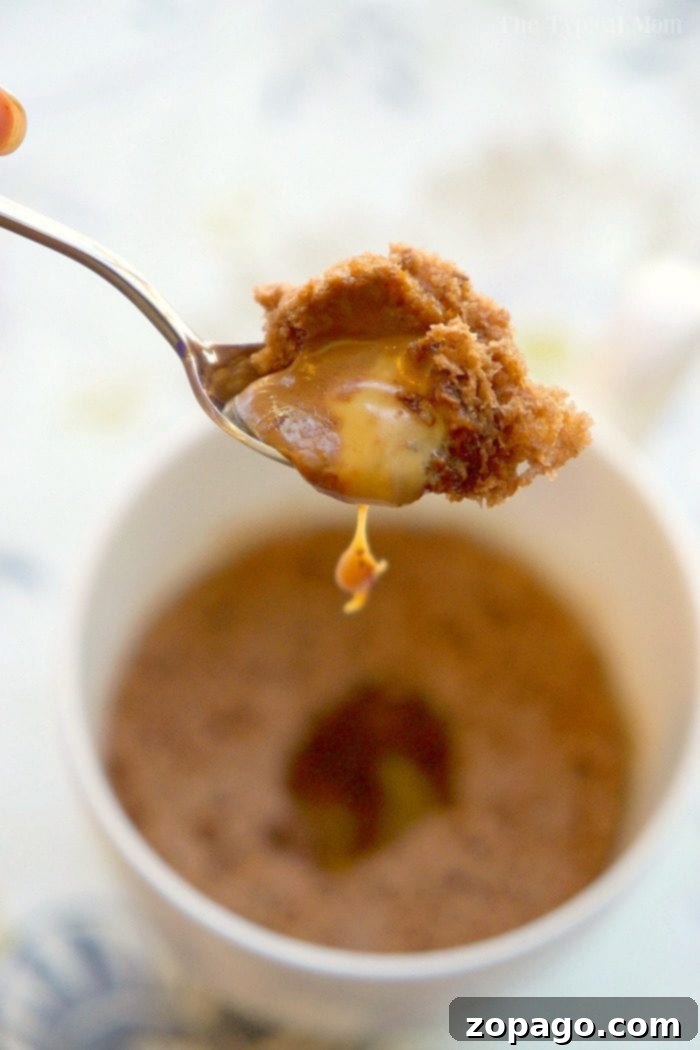 Gooey Caramel Chocolate Mug Cake A spoon lifts a decadent bite of molten caramel and chocolate mug cake, revealing a gooey center within a white mug.