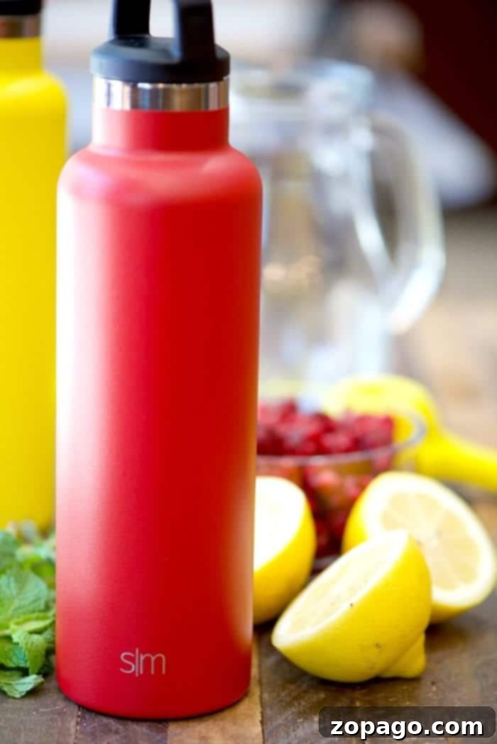 This homemade raspberry lemonade recipe only requires 4 ingredients and is perfect for Summertime! Packed with fresh fruit it's my favorite on the go drink. #homemade #raspberry #lemonade #drink #juice A vibrant red insulated water bottle surrounded by fresh lemons and a pitcher of homemade raspberry lemonade, highlighting refreshing ingredients.