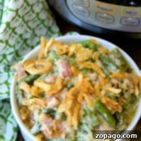 A bowl of instant pot green bean casserole topped with crispy onions sits beautifully in front of a pressure cooker, with a green patterned cloth nearby.