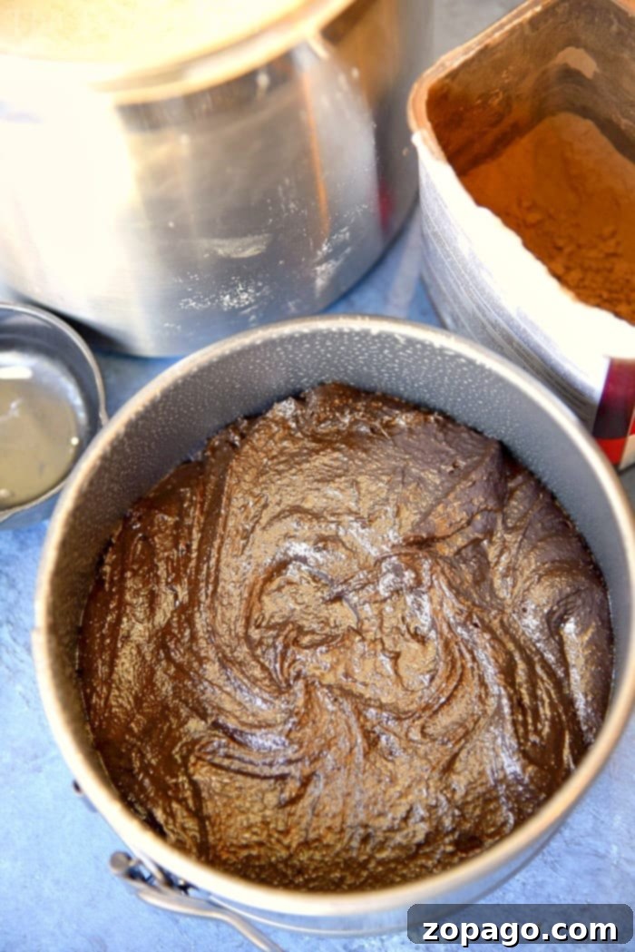 This easy Instant Pot chocolate cake with raspberry sauce recipe is so amazing you won't want to share! The moistest cake ever and only takes 35 minutes! #instantpot #pressurecooker #chocolate #cake #dessert Cake batter in a round baking pan sits next to a bowl and a box of cocoa powder on a blue countertop, waiting to transform into an instant pot chocolate cake.