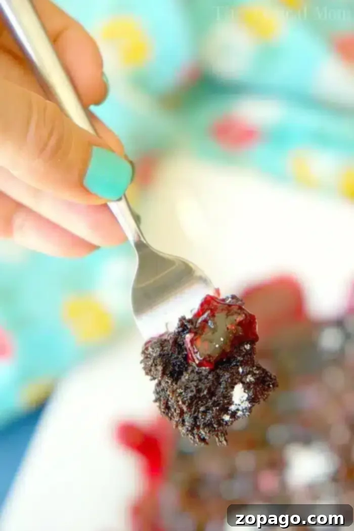Instant Pot Chocolate Cake Perfection 3 A hand with teal-painted nails holds a fork with a bite of instant pot chocolate cake topped with red berry sauce. In the background, a blurred colorful tablecloth is visible with more of the dessert on a plate, inviting another taste.
