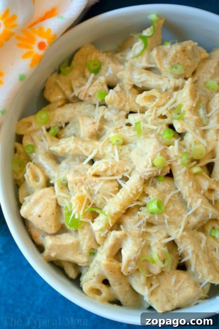 This easy crockpot pesto chicken alfredo casserole is a hit in my house! Cheesy noodles with moist pieces of pesto chicken together makes a complete meal. Serving of crockpot pesto chicken alfredo casserole on a plate, showing the creamy pasta, chicken pieces, and a garnish of fresh green onions.