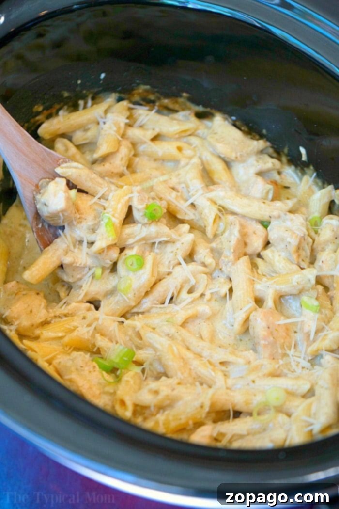 This easy crockpot pesto chicken alfredo casserole is a hit in my house! Cheesy noodles with moist pieces of pesto chicken together makes a complete meal. Overhead shot of crockpot pesto chicken alfredo casserole with a serving spoon, garnished with fresh basil, ready to be enjoyed.