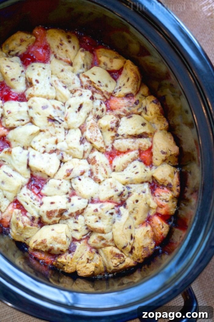 Slow Cooker Cinnamon Roll Pull-Apart Bread 2 A close-up view of a decadent dessert in a slow cooker. The dish features golden-brown biscuit pieces reminiscent of cinnamon roll monkey bread, resting on a red, fruit-filled base, likely a cobbler. The crockpot has a black rim and sits on a burlap surface.
