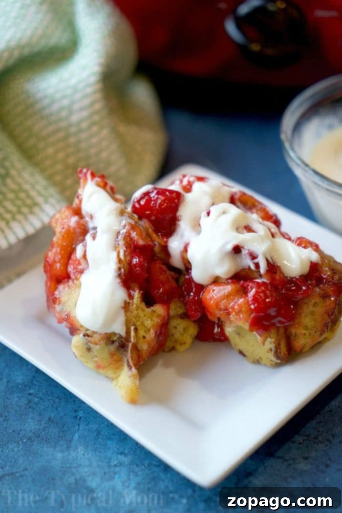 This easy crockpot cinnamon roll monkey bread casserole is perfect for dessert or breakfast! Use any pie filling you like and with 3 ingredients it's done! A white plate with two pieces of French toast drizzled in strawberry sauce and cream rests on a blue surface, evoking the coziness of crockpot cinnamon roll monkey bread. In the background, a green and white cloth accompanies a small bowl of additional cream.