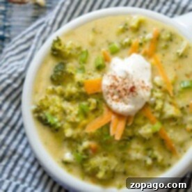 Bowl of quick broccoli cheese soup topped with shredded carrots, green onions, and a dollop of sour cream on a striped cloth.