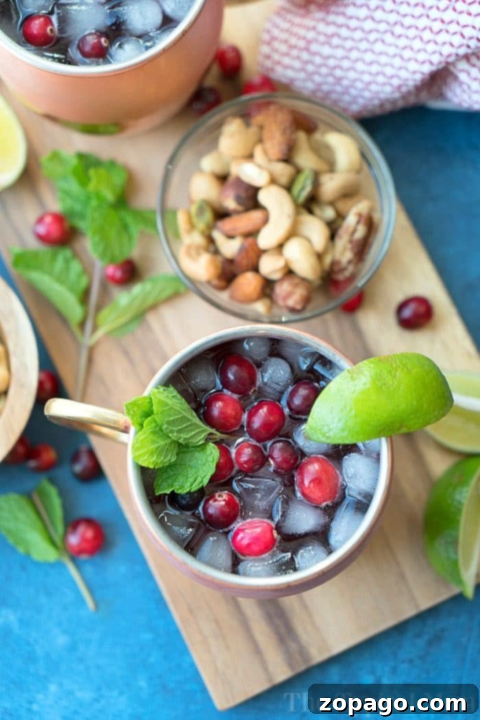 Serve up holiday cheer with this beautiful Cranberry Moscow Mule, a treat for both eyes and palate. A glistening copper mug filled with a Cranberry Moscow Mule mocktail, adorned with fresh cranberries, a vibrant mint sprig, and a lime wedge. A rustic wooden board with a bowl of nuts sits nearby.