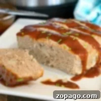A sliced turkey meatloaf topped with a glossy brown sauce and garnished with chopped green onions sits on a white square plate. The Instant Pot, which made this tender loaf possible, is visible in the blurred background alongside a blue cloth.