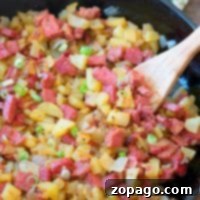 A skillet containing a homemade corned beef hash recipe with diced corned beef, potatoes, onions, and green onions is being stirred with a wooden spoon.