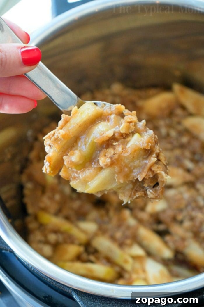 Instant Pot apple crisp recipe is amazing! Tastes like copycat Cracker Barrel baked apples we love but made in less than 20 minutes total. Warm cinnamon apples coated with a ooey gooey brown sugar glaze your family will go crazy over for sure. Try this homemade pressure cooker fruit dessert this week! #instantpot #pressurecooker #apples #applecrisp #dessert #bakedapples #instantpotrecipes #thetypicalmom #easy A hand holding a spoonful of crumbly apple crisp over an Instant Pot filled with delicious apple crisp.