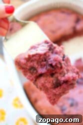 A close-up of a person's hand holding a spatula with a cherry pie cake slice made from cherry pie filling. The cake has a fluffy texture, containing dark, maroon-colored fruit bits within it. A pan with the remaining dump cake is in the background.