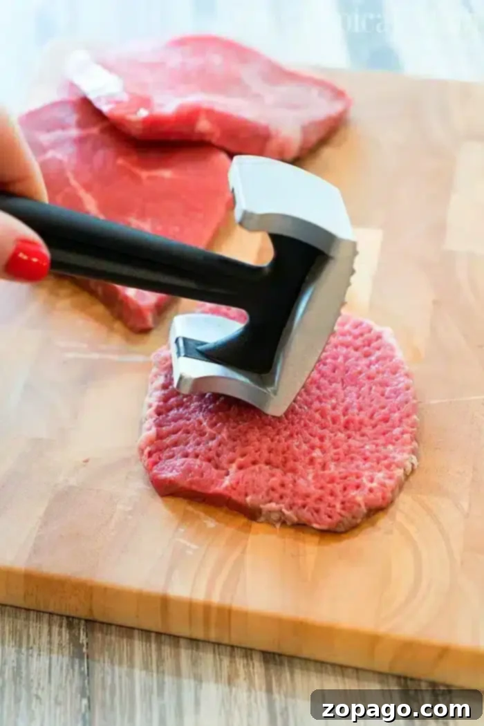 Instant Pot cube steak with gravy is yummy! Tender bites of meat smothered in a creamy mushroom sauce. A total winner. #instantpot #pressurecooker #cubesteak #steak #thetypicalmom #instant-pot-recipes A person uses a meat tenderizer to pound a slice of raw beef on a wooden cutting board, preparing it for a Ninja Foodi cube steak recipe. Two more slices of raw beef are visible in the background on the cutting board.