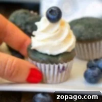 A hand with red nail polish holds a mini blueberry cupcake adorned with white frosting and a fresh blueberry on top. Other cupcakes, scattered blueberries, and hints of blueberry pie filling create a tempting backdrop on a white plate and wooden surface.