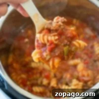 A hand holds a white ladle filled with Instant Pot sloppy joe casserole—rotini pasta, ground meat, and rich tomato sauce—over an open pressure cooker brimming with the same delicious mixture.