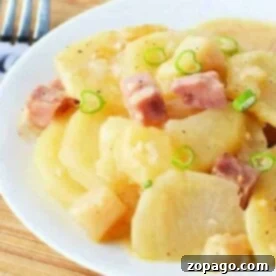 A plate of slow cooker scalloped potatoes topped with diced ham and green onions sits invitingly on a wooden board, accompanied by a fork.