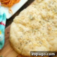 Homemade slow cooker rolls topped with herbs and shredded cheese sits on a wooden surface, while slow cooker rolls cool beside it. In the background, a corner of a white plate with spaghetti can be seen, complemented by a colorful cloth and a sprig of fresh herbs nearby.