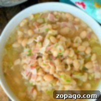 A bowl of pressure cooker bean soup with bacon bits, garnished with herbs, sits next to a patterned cloth.