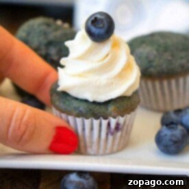 A hand with red nails holds a blueberry cupcake, elegantly topped with cream and a blueberry—a delightful treat straight from an easy recipe with blueberries.