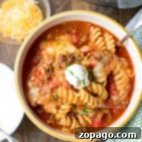 A bowl of cheesy taco pasta with rotini, topped with sour cream, sits enticingly next to a tempting bowl of shredded cheese and the very Instant Pot that crafted this delightful instant pot taco casserole.