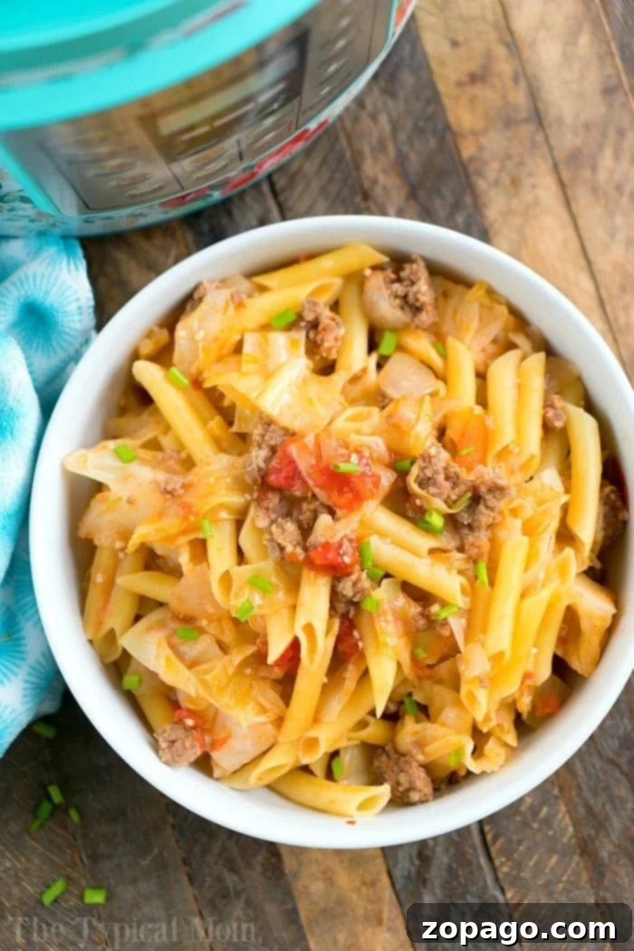 Instant Pot Sausage and Cabbage Casserole with Noodles A bowl filled with penne pasta, perfectly cooked in a pressure cooker, mixed with ground beef, chopped tomatoes, and savory sausage. The dish is topped with green onions and accompanied by crisp cabbage on a wooden table beside a blue patterned cloth.