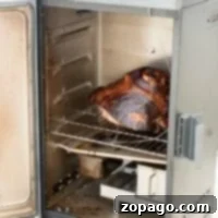 A metal smoker with the door open reveals a large smoked cooked ham on the rack inside. The smoker rests on a wooden surface.