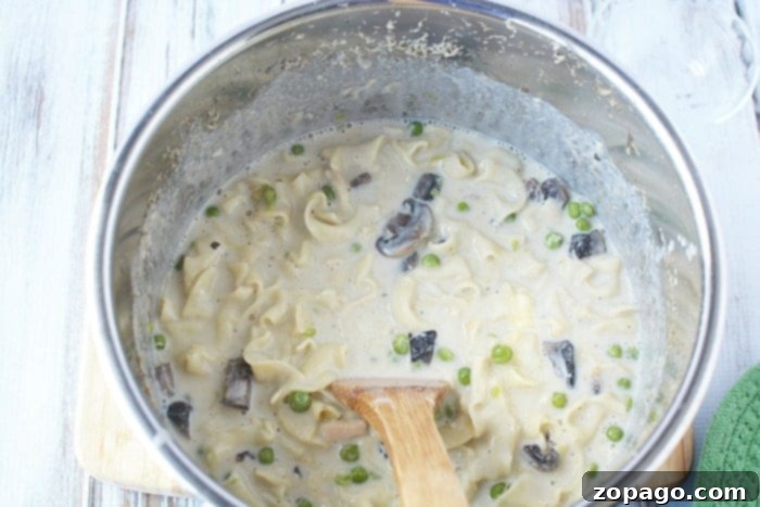 This pressure cooker tuna noodle casserole will bring you back to your childhood for sure. The comfort food you love can now be made in your Instant Pot for dinner in no time at all and is loaded with all the ingredients you once made in your oven. If you're looking for a new Instant Pot recipe to try this is it! #instantpot #pressurecooker #tunanoodlecasserole #casserole #tuna #pasta #dinner A pot of creamy tuna noodle pasta with mushrooms and peas, reminiscent of a classic pressure cooker tuna noodle casserole, is gently stirred with a wooden spoon on a rustic wooden surface.