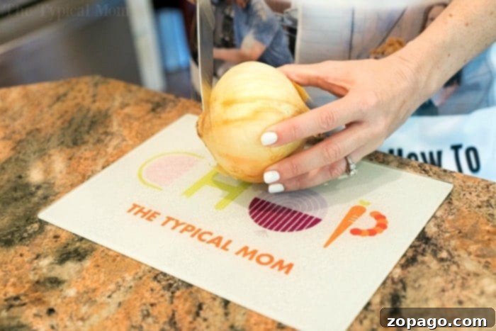 Instant Pot Keto Cheeseburger Soup 4 A person with white nail polish is slicing an onion on a cutting board marked with "CHOP THE TYPICAL MOM," preparing ingredients for an instant pot cheeseburger soup. The cutting board rests elegantly on a granite countertop.