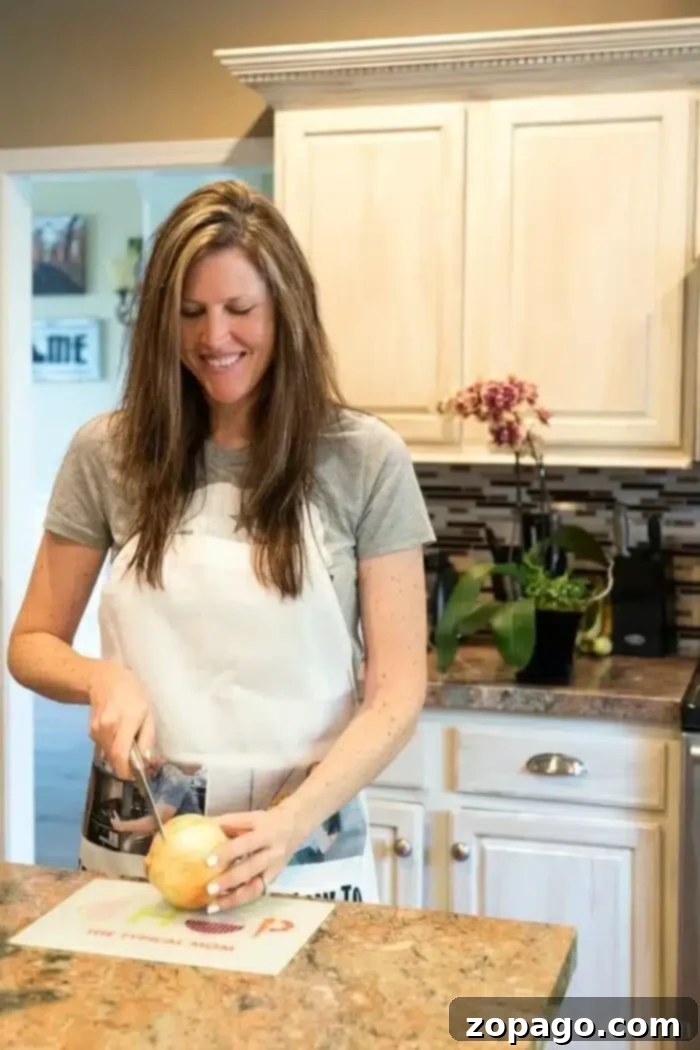 Instant Pot Keto Cheeseburger Soup 5 A woman with long hair stands in the kitchen, smiling as she slices a large onion on a cutting board. Dressed in a gray t-shirt and white apron, she's preparing ingredients for an instant pot cheeseburger soup. Plants and kitchen items line the countertop behind her.