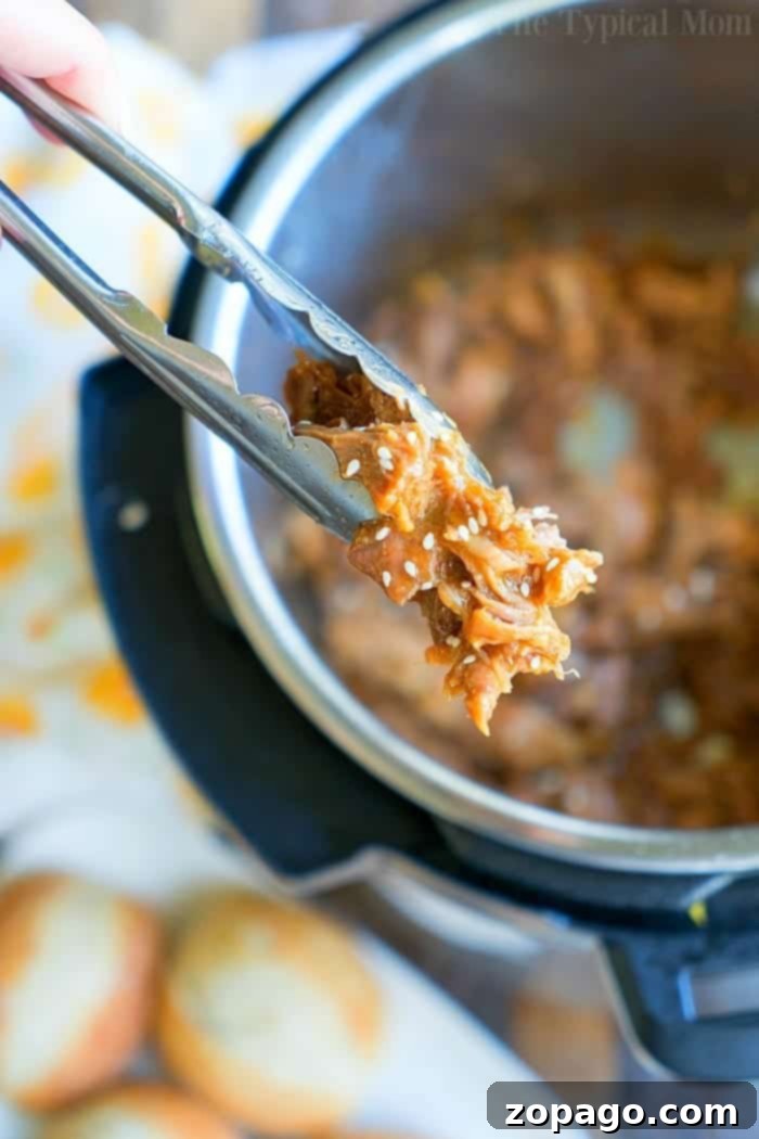 Asian Instant Pot pulled pork recipe that's sticky sweet with a little kick! Tender pork roast in an hour using your pressure cooker. The best sliders ever! #instantpot #instantpotrecipes #pressurecooker #pork #pulledpork #honey #sliders #appetizer #asian Tongs hold a portion of teriyaki pulled pork over an open Instant Pot. The succulent meat is mixed with sauce and sprinkled with sesame seeds. A blurred background reveals bread rolls and part of a printed cloth, adding a cozy touch to this culinary scene.