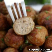 A fork holds a halved meatball above a pile of perfectly cooked, flavorful spheres, garnished with fresh herbs. These delectable bites are straight from frozen meatballs in an air fryer for that crispy perfection.