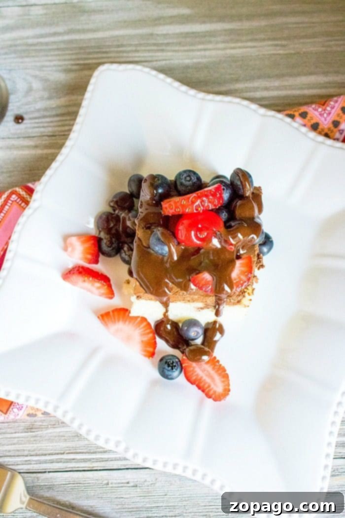 A slice of poke cake topped with chocolate sauce, fresh strawberries, and blueberries on a white plate, inviting you to learn how to make a poke cake for your next celebration.