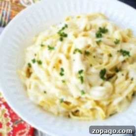 A white bowl filled with creamy Instant Pot fettuccine alfredo, garnished with chopped parsley. The bowl is placed on a colorful patterned tablecloth, with a fork nearby.