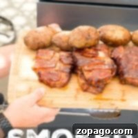 A person holds a wooden tray laden with bacon-wrapped smoked pork chops and baked potatoes by a grill.