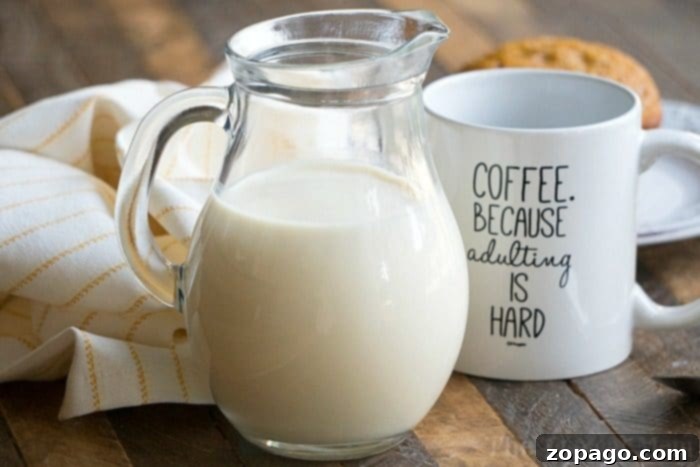 A glass pitcher filled with pumpkin spice coffee creamer sits beside a coffee mug, emblazoned with 'COFFEE. BECAUSE ADULTING IS HARD', on a rustic wooden table, evoking cozy autumn vibes.