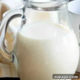 A clear glass pitcher filled with milk and pumpkin spice coffee creamer sits on a wooden table next to a white cloth.