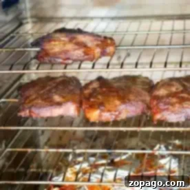 Four pieces of smoked pork steaks with dry rub are placed on two smoker racks, with a foil-lined tray catching drippings below. The meat appears perfectly browned, indicating it has been roasted or broiled to smoky perfection.