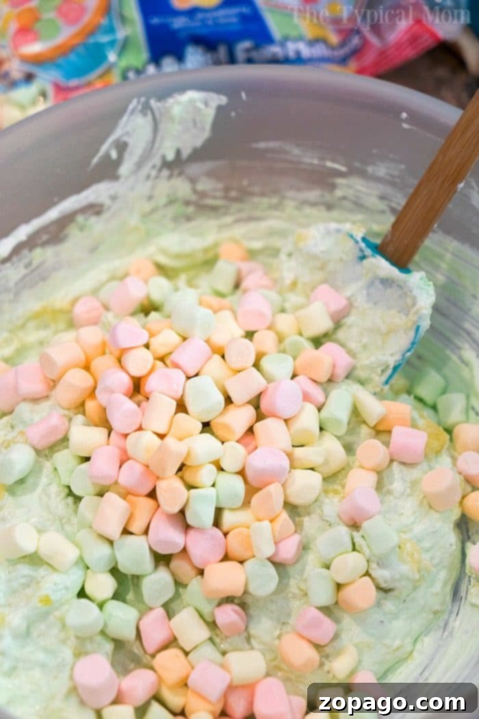 Pistachio pudding ambrosia is an amazing side dish or dessert! A cross between your favorite pistachio pudding and marshmallow ambrosia, you'll love it too. #pistachiopudding #ambrosia #dessert #marshmallow #pistachio #sidedish #thanksgiving Close-up of Pistachio Pudding Ambrosia with mini marshmallows, pineapple, and coconut, served in a glass bowl.